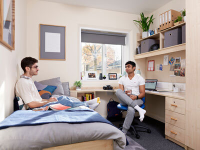 Student sitting on the bed while another sits on a chair in student accommodation chatting.