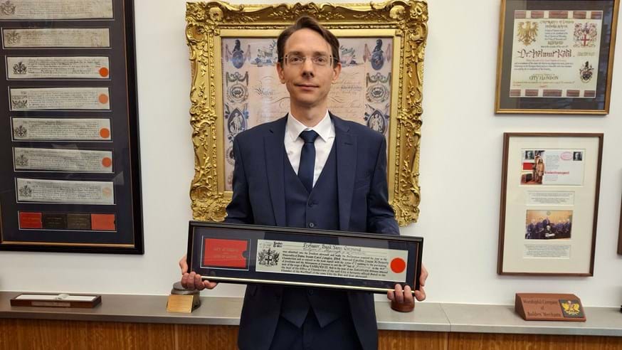 Image shows Professor Germond with a framed copy of the Freedom at Guildhall against a backdrop of other framed items