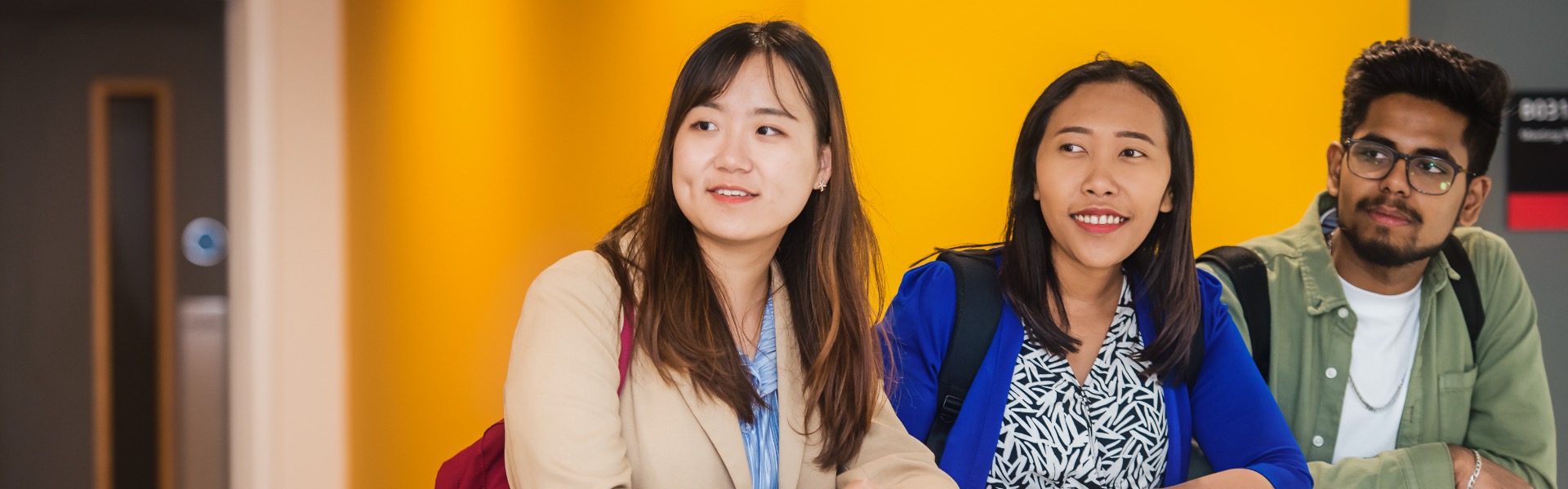Three students lean on a balcony against a bright yellow wall