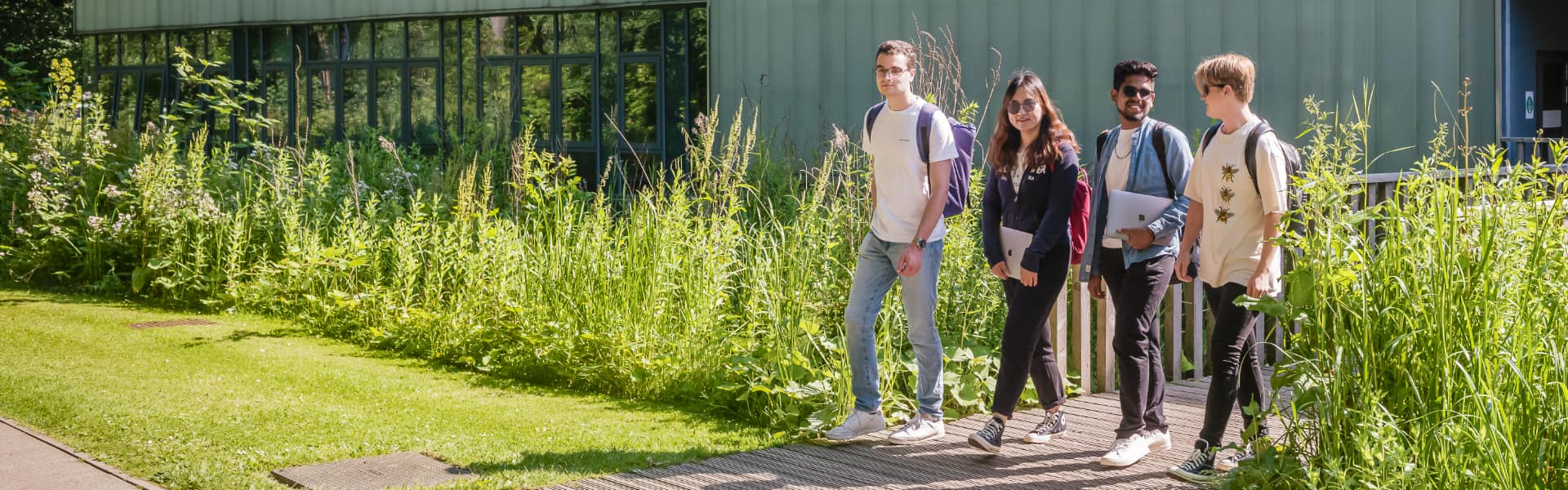 Four students walk across a wooden bridge, leaving a modernist green building.