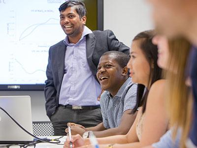 Lecturer standing next to a group of students who are sitting making notes and smiling.