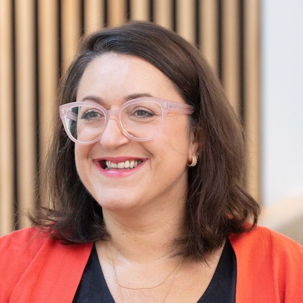 A female wearing glasses with dark hair, standing in front of a wooden wall.