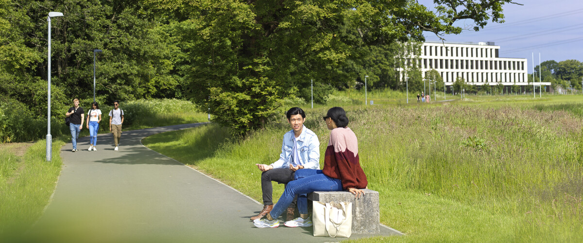 Students sit on a bench in the campus parkland. More people walk along a path in the background.