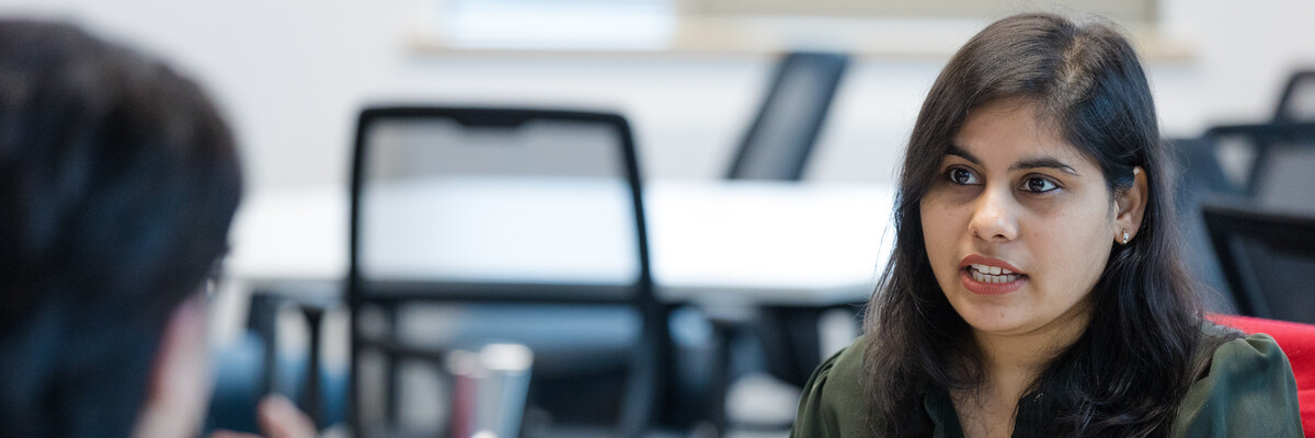 A person sat behind a laptop talks to someone, out of focus, over a desk.