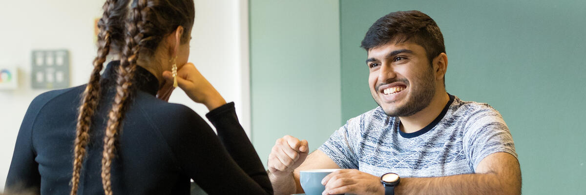 A student smiles while holding a cup of coffee and talking to someone on the other side of the table